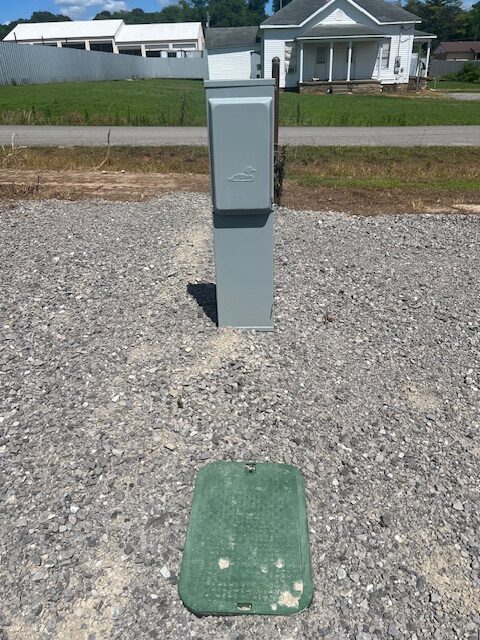 A gray utility box stands on a gravel surface with a green rectangular cover lying flat in front of it. There is grass and a road in the background, along with a white house and other buildings.