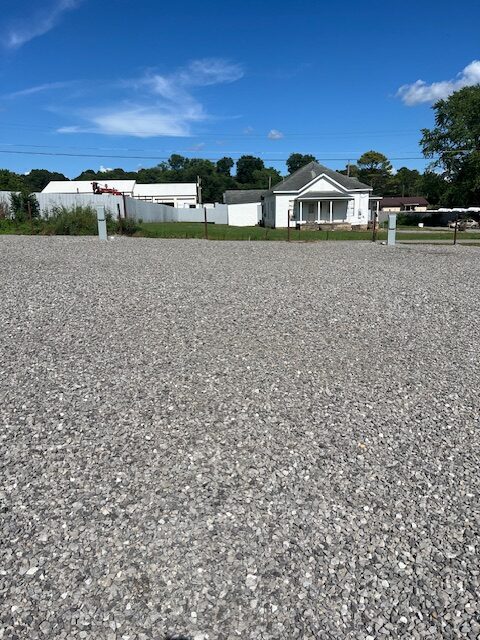 A gravel lot stretches across the foreground, with a small white house, a fence, and trees visible in the background under a clear blue sky.