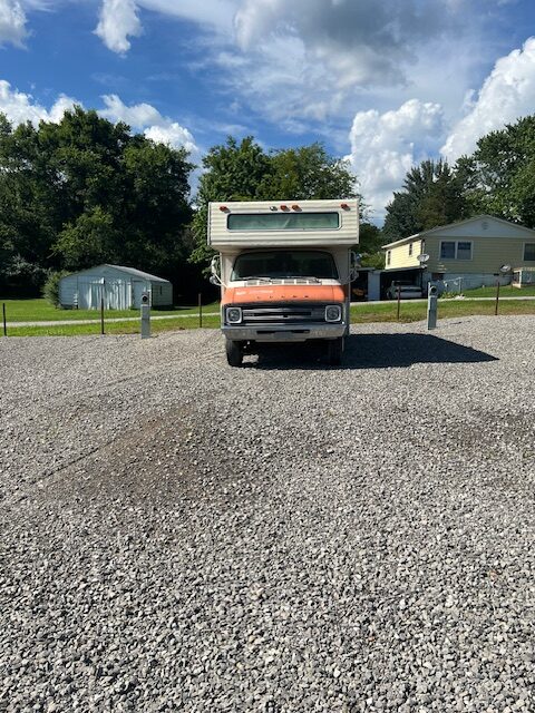 A vintage orange and white RV is parked on a gravel lot under a partly cloudy sky, with houses, trees, and utility posts visible in the background.