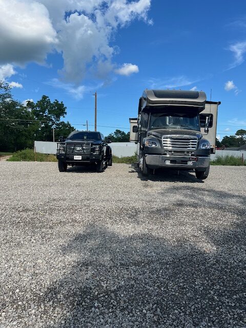 A black SUV and a large gray RV are parked side by side on a gravel lot under a blue sky with scattered clouds. Trees and a fence are visible in the background.